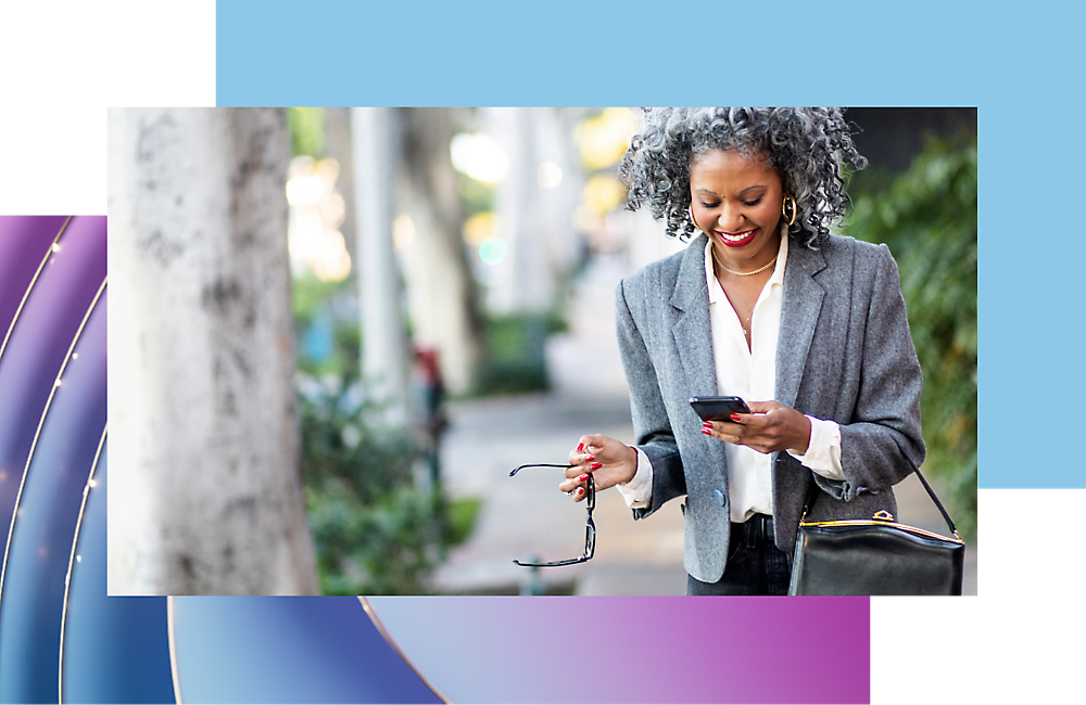 A smiling woman with gray curly hair looking at her smartphone and holding glasses, stands on a city sidewalk.