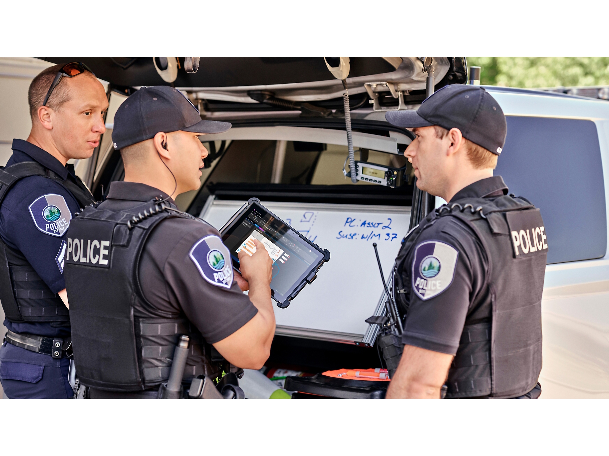 Three police officers review information on a digital tablet beside their patrol car with the trunk open