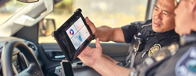 Two police officers in a vehicle, one holding a tablet displaying a map, engaged in a discussion.