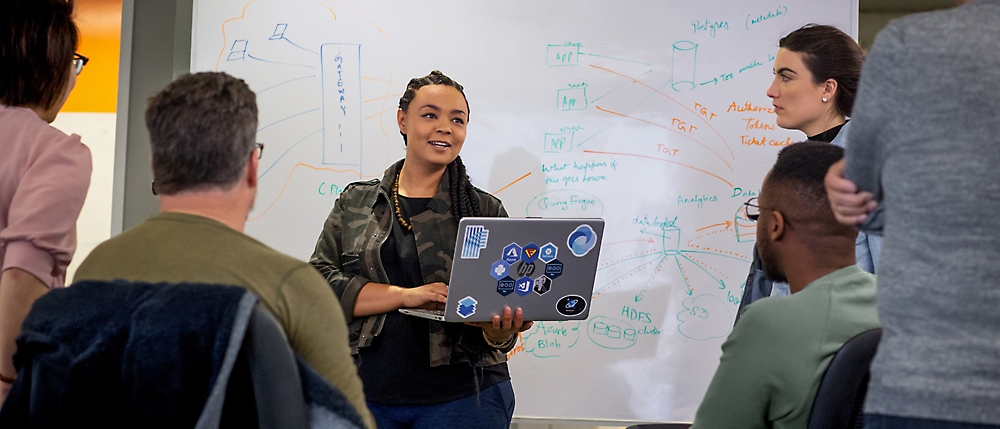 A woman presents a business strategy on a clipboard during a meeting with colleagues in front of a whiteboard.