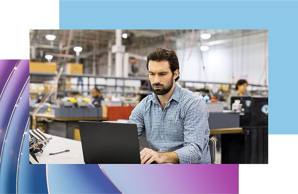 Um homem a trabalhar num computador portátil num ambiente industrial com maquinaria e trabalhadores em segundo plano.