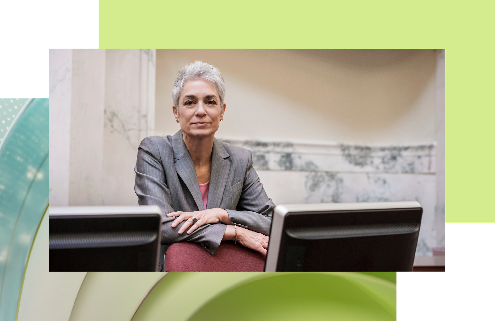 A professional woman with short grey hair, wearing a grey blazer, sitting at a desk with laptop computers