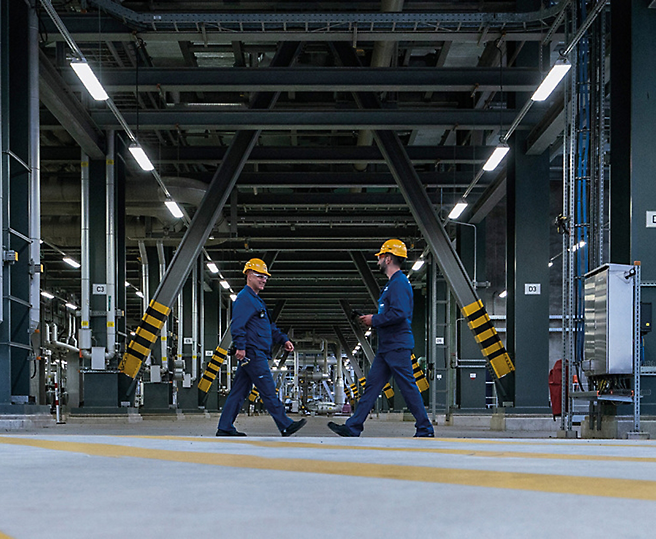 Dois trabalhadores com capacetes de proteção a caminhar numa instalação industrial.