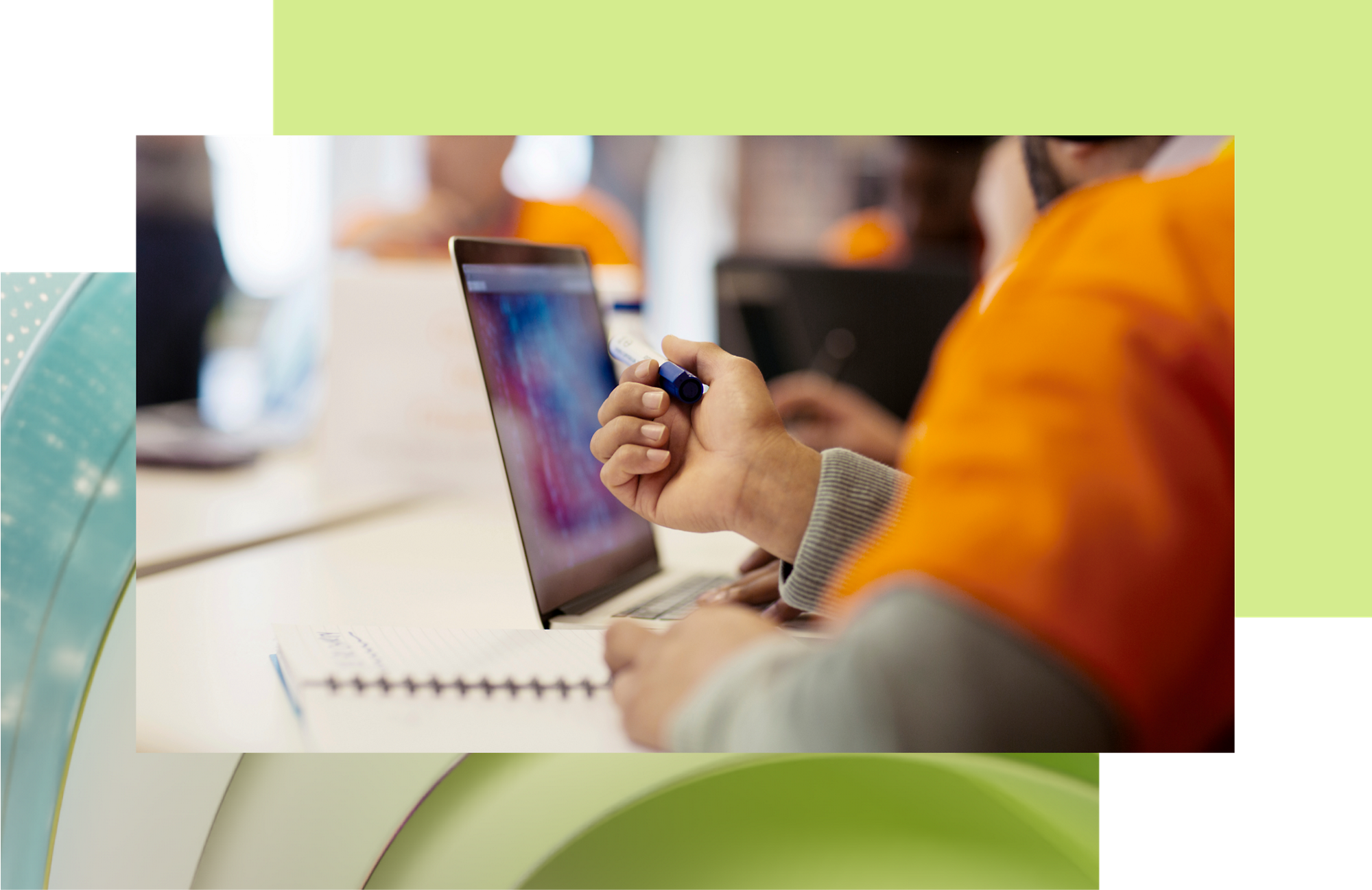 A person in an orange jacket using a stylus on a laptop screen at a busy workspace.