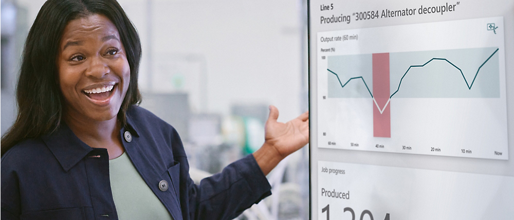 A woman smiles while presenting a productivity graph on a digital display in an industrial setting.