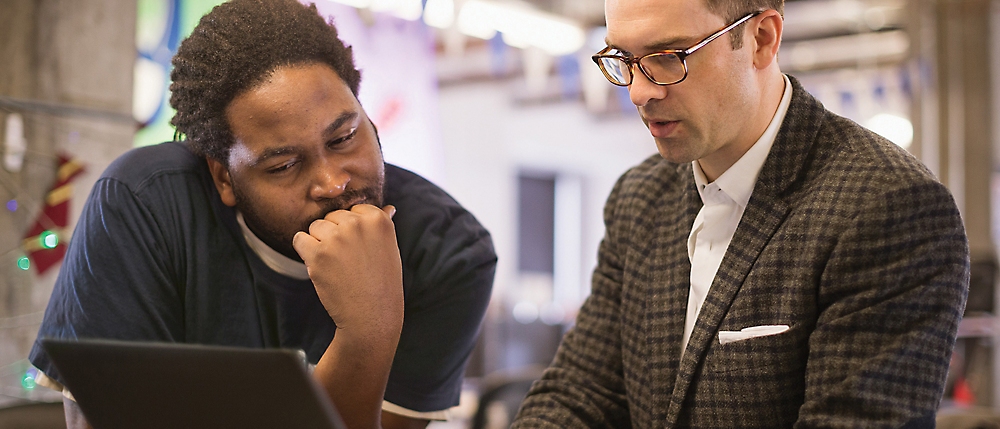 Two men, one african american and one caucasian, intently review content on a laptop in an office setting.