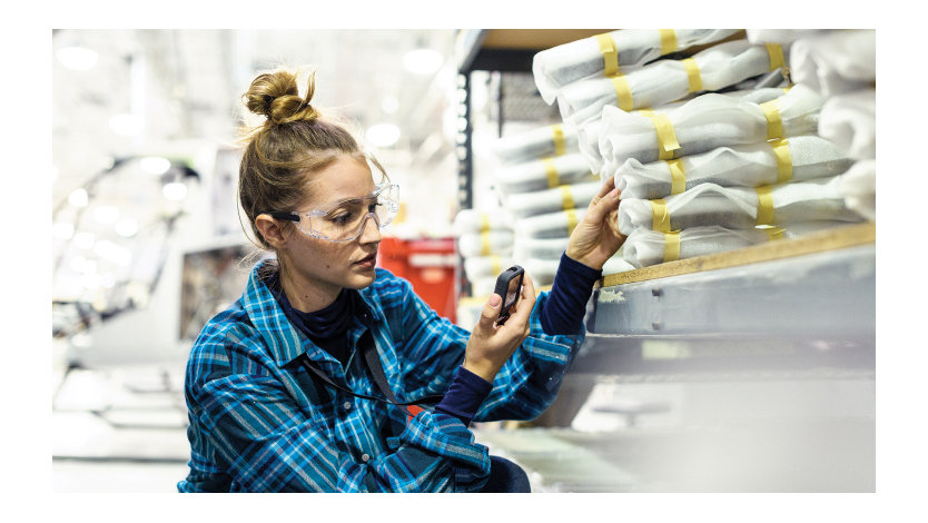 A female worker wearing safety glasses using a device kneels in front of products on shelving in a manufacturing storage space.