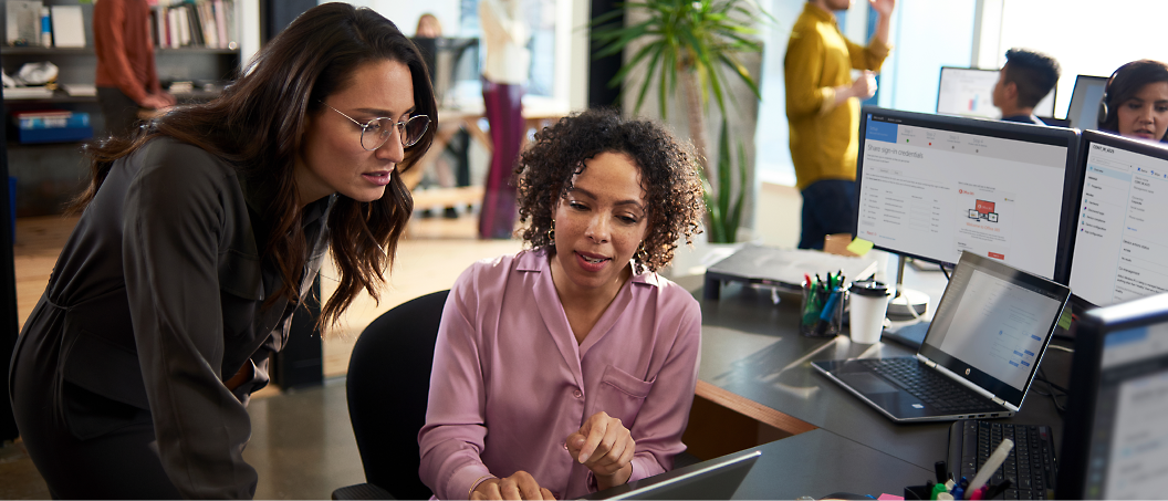 Two women discussing work in front of a laptop in a busy office setting.