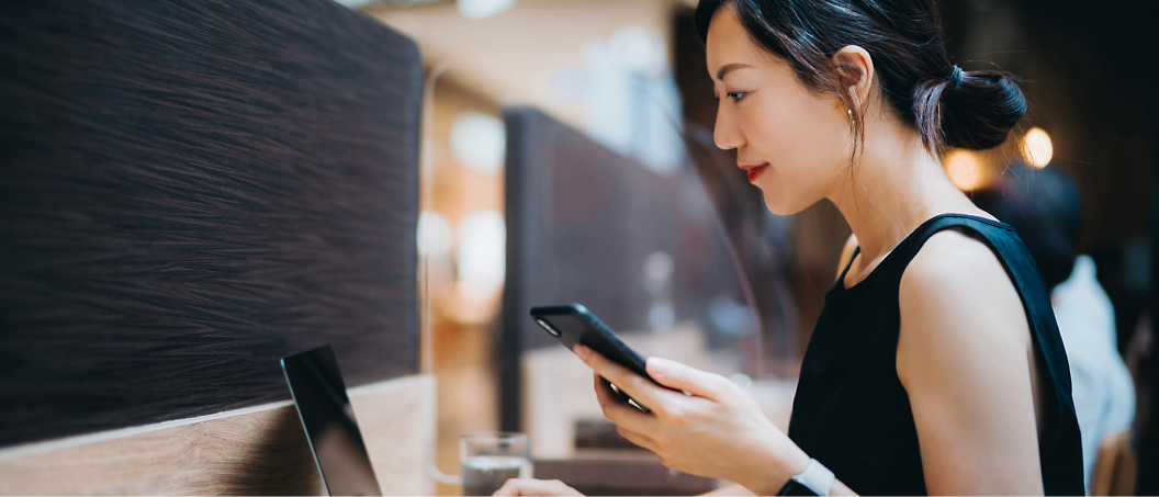 Woman in a black dress using a smartphone while sitting at a café table near a window.