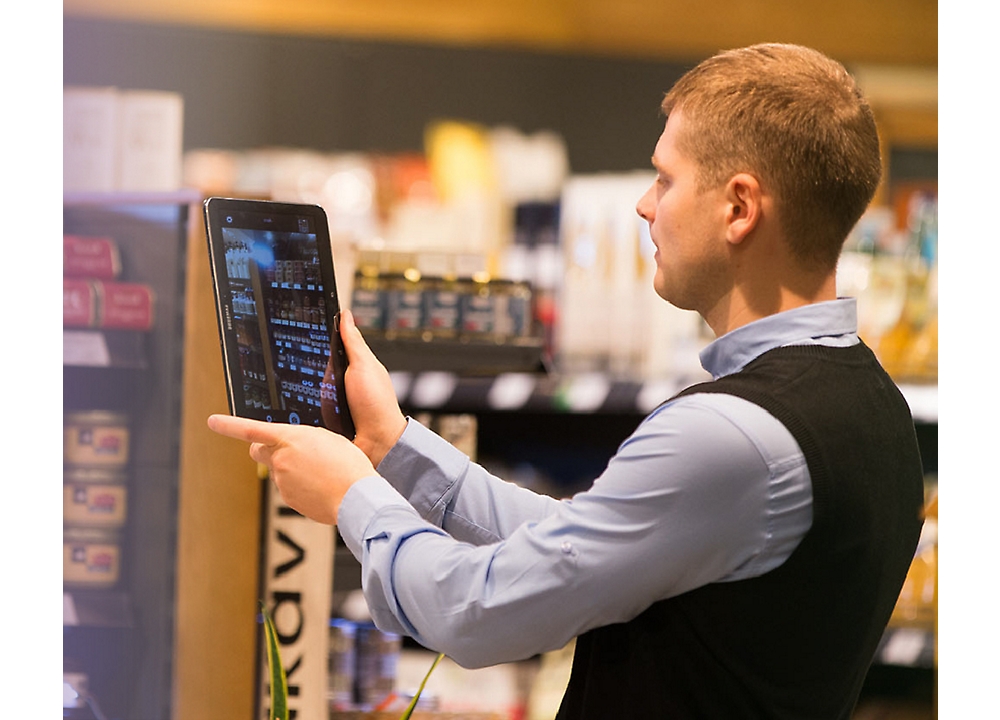 A person holds a tablet, taking inventory in a store aisle, with various products visible on the shelves in the background.