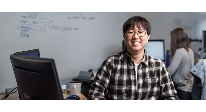 화이트보드가 있는 사무실의 컴퓨터 앞에 앉은 안경과 체크무늬 셔츠를 입은 미소 짓는 동아시아 사람 