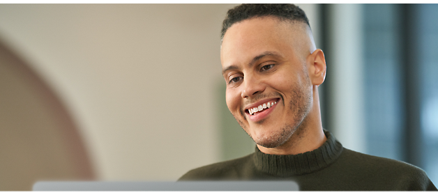 A man wearing a dark green sweater is smiling while looking at a laptop screen.