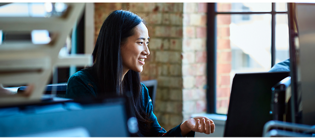 A woman with long black hair sits at a desk, smiling and gesturing with her hand