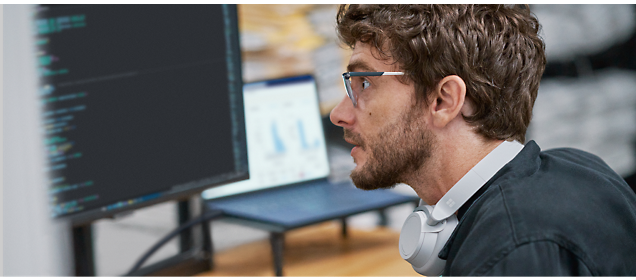 Man wearing glasses and headphones looks at computer screen displaying code, with a laptop in the background.