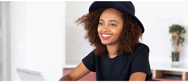 A woman with curly hair wearing a dark blue hat and black shirt smiles while sitting at a table with a laptop.