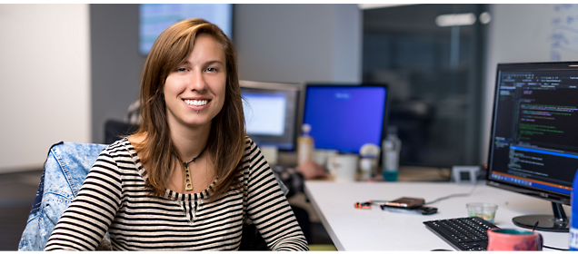 A woman with long brown hair, wearing a striped shirt, smiles while sitting at a desk with computer monitors 