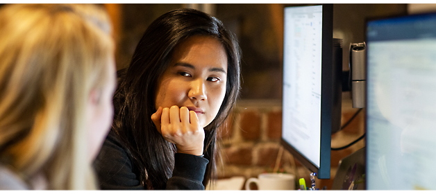 Two people are conversing at a workspace, with one person looking intently at the other while resting their chin on their hand