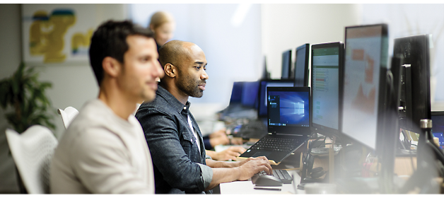 Several people are working at desks with computers in an office environment. The focus is on two men typing on their keyboards.