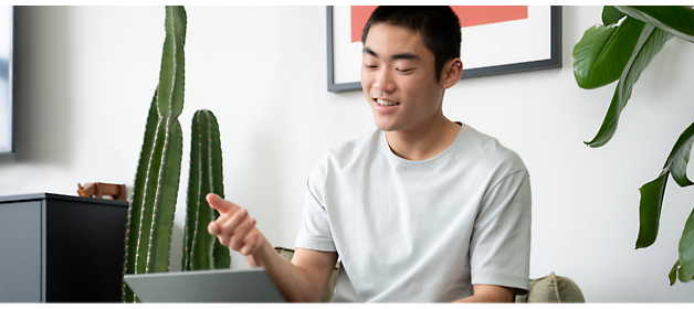 A person in a light gray shirt sits indoors, smiling and gesturing while using a laptop