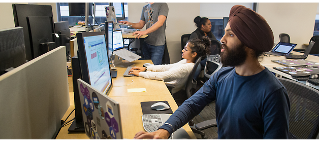 Four people in an office work at their computers. One stands at a standing desk, while the others sit