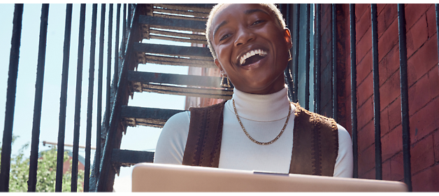 A person is sitting outdoors on a staircase, smiling, wearing a white turtleneck and a brown vest, and working on a laptop.