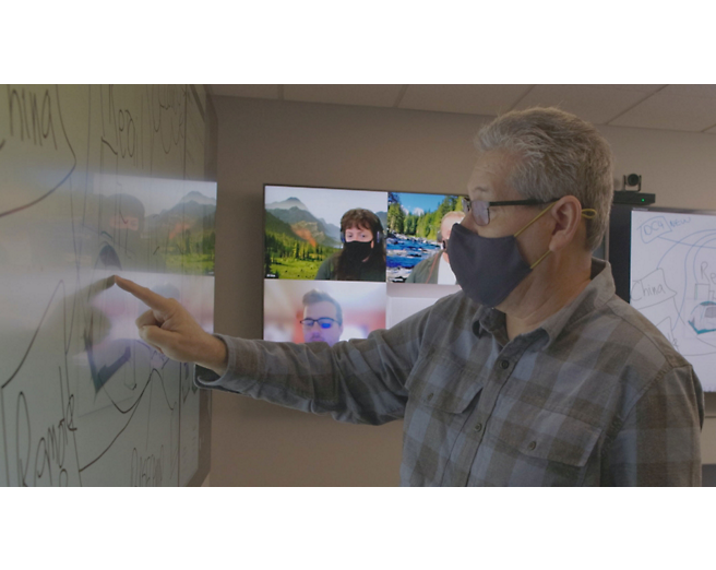 A man wearing a face mask is pointing at a screen.