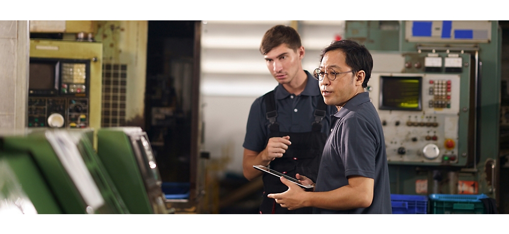 Two people looking at some machine and in that one person is wearing glasses and holding tablet.