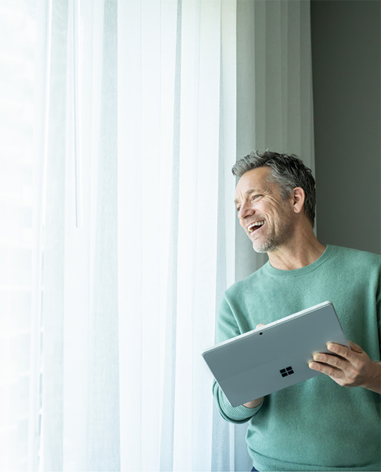 A man standing, smiling and looking out a window while holding a Surface device