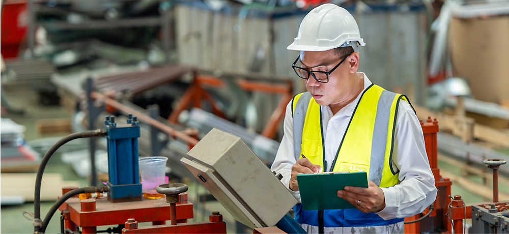 A worker wearing a white hard hat, safety glasses, and a yellow reflective vest records data on a clipboard in an industrial factory setting with machinery and equipment in the background.