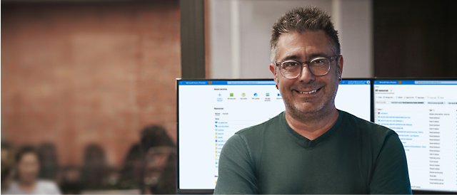 A person wearing spectacles smiling with a desktop screen in the background.