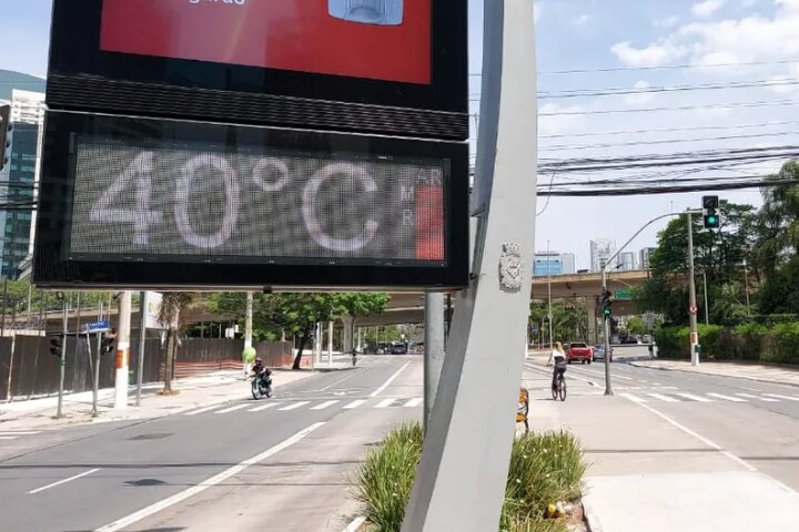 Na imagem, vê-se um termômetro que marca 40 graus celsius de temperatura, localizado na avenida Berrini. Na via à esquerda vemos um motociclista, enquanto, na via à direita, vemos uma ciclista, uma caminhonete vermelha, um carro prateado e um farol com luzes acesas de cor verde.