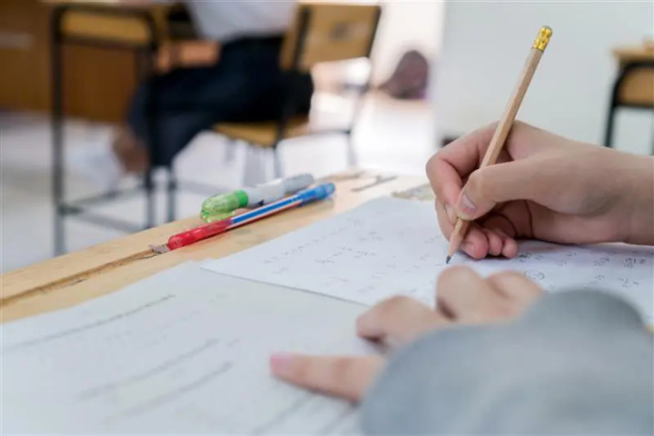 Na imagem vê-se a parte de cima de uma carteira escolar, com as mãos de um aluno apoiadas sobre ela, segurando um lápis na mão direita resolvendo exercícios de matemática e duas canetas a sua frente, na borda da mesa.