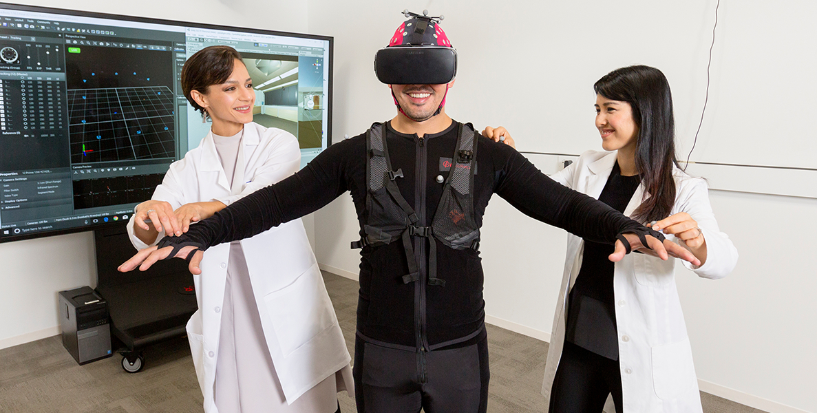 Two female researchers in lab coats working with male wearing advanced virtual reality headset