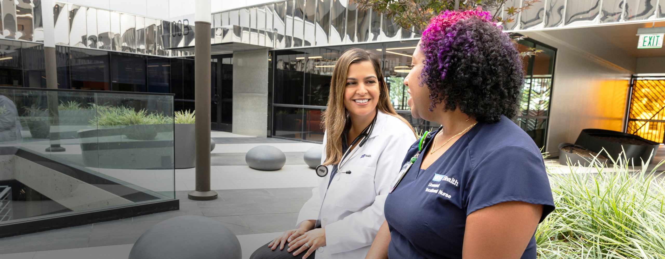 A smiling female UCLA Health physician and a smiling female UCLA Health nurse talking together in a courtyard outside of a UCLA Health hospital