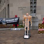 Protesters hold a sign that says, "Sacklers are murderers," and display a "Pill Man" sculpture that consists of pill bottles assembled into the shape a human skeleton.