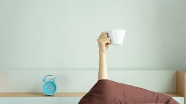 Woman showing arm raised up holding coffee cup on bed under duvet in the bed room, Young girl with hand sticking out from the blanket holding tea cup. wake up drinking coffee in morning concept.