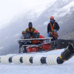 Image of two humans sitting behind a control console dressed in heavy clothing, while a long tube sits on the ice in front of them.