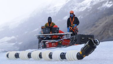 Image of two humans sitting behind a control console dressed in heavy clothing, while a long tube sits on the ice in front of them.