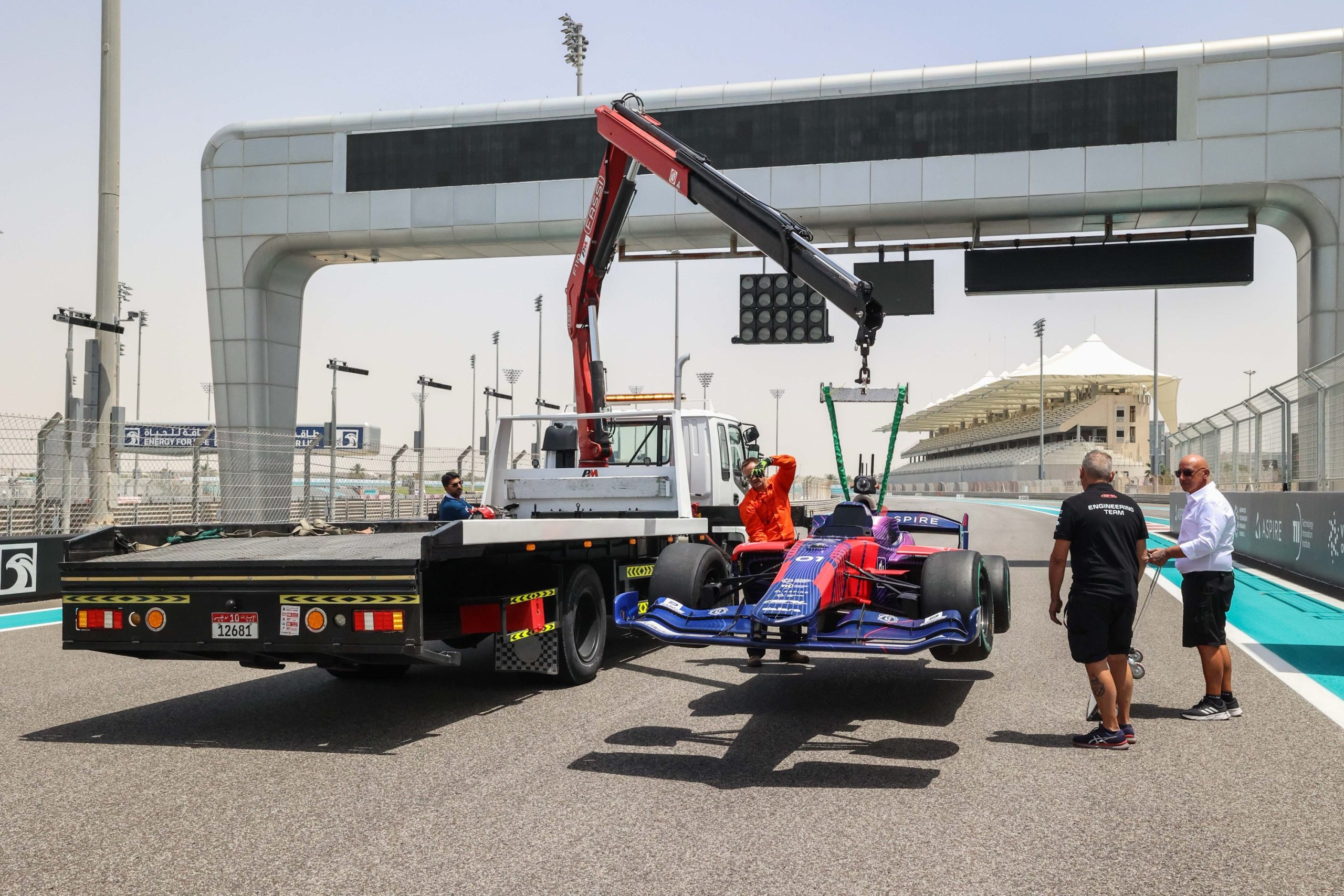 A blue and red Dallara SF23 race car is lifted by a crane attached to a flatbed truck.