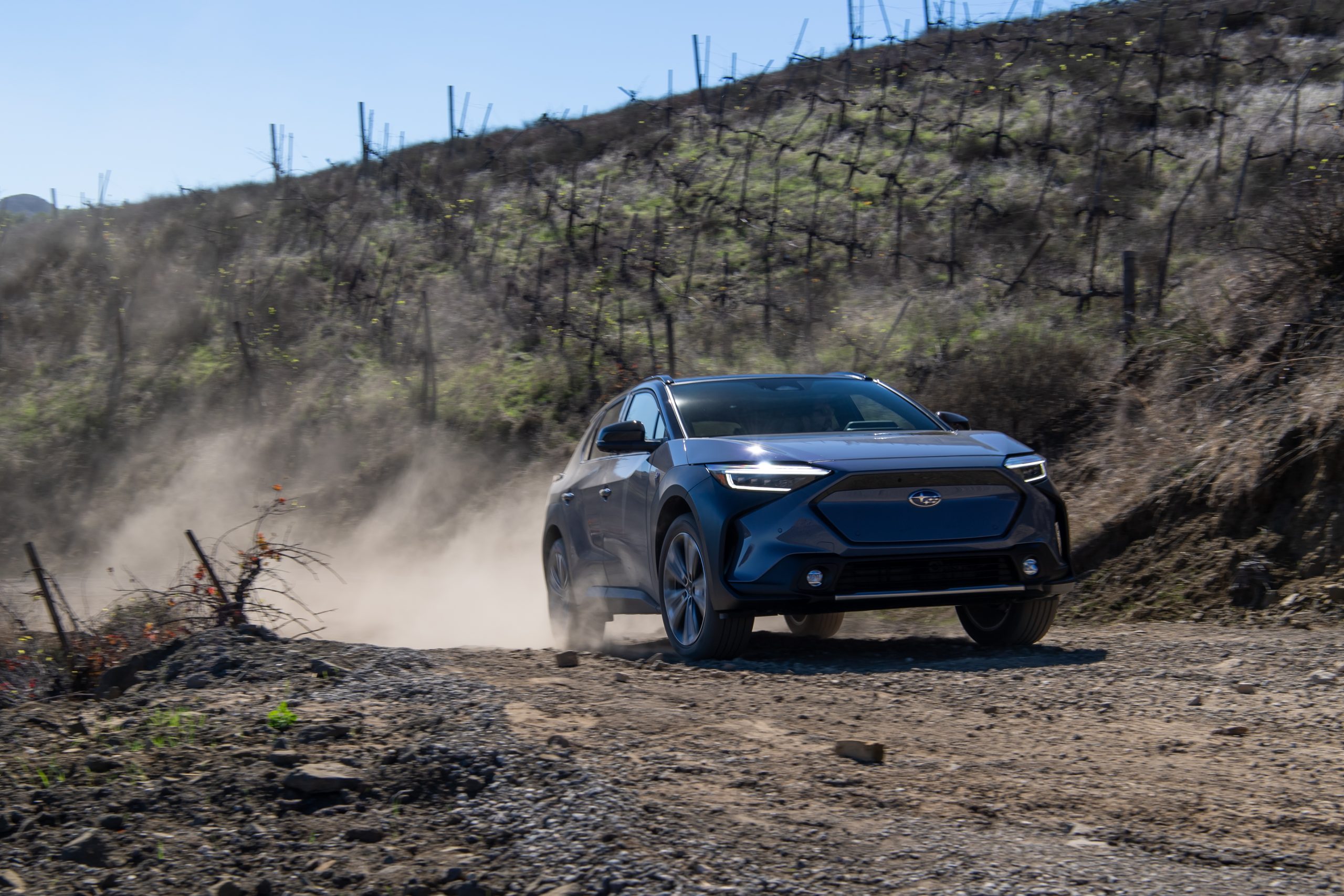 A Subaru Solterra drives on a dirt road past a vineyard