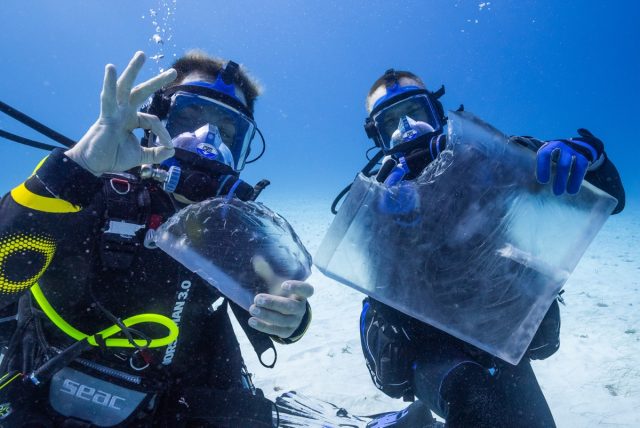 Two men in scuba gear underwater, one on left gives thumbs up as one on right holds gelatin cube with a bite off the corner.