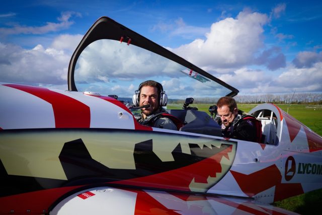 two men in the cockpit of a small aircraft painted red and yellow with black markings