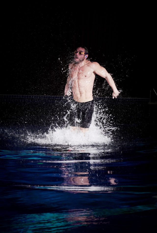 well muscled man in capri speedos emerging in slow motion out of a pool, 3/4 of his body clearing the surface.