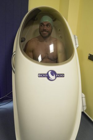 well muscled man in a human-sized white cylinder with a large window in front through which he can see