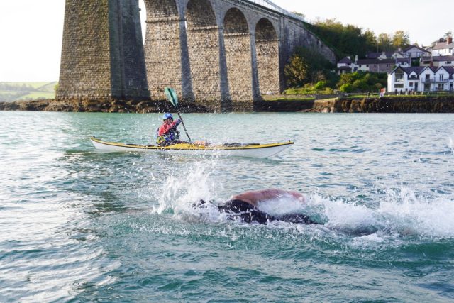 man swimming in open water while a kayaker monitors him in the background