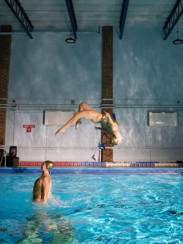 female synchronized swimmer flying through the air horizontally with arched body after being propelled from the water by a teammate