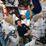 Image of two astronauts in an equipment filled chamber, standing near the suits they wear for extravehicular activities.