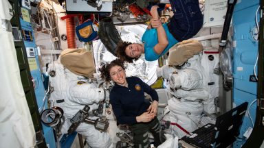 Image of two astronauts in an equipment filled chamber, standing near the suits they wear for extravehicular activities.