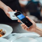 A raised iPhone-ish phone taps against a mobile payments square above restaurant dishes, including prawns.