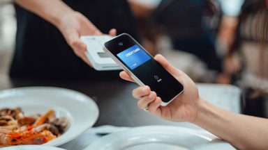 A raised iPhone-ish phone taps against a mobile payments square above restaurant dishes, including prawns.